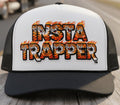 A black and white trucker hat featuring a white front panel with the text 'INSTA TRAPPER' in bold black letters filled with orange and yellow tiger illustrations and flames. The brim and mesh back are black.